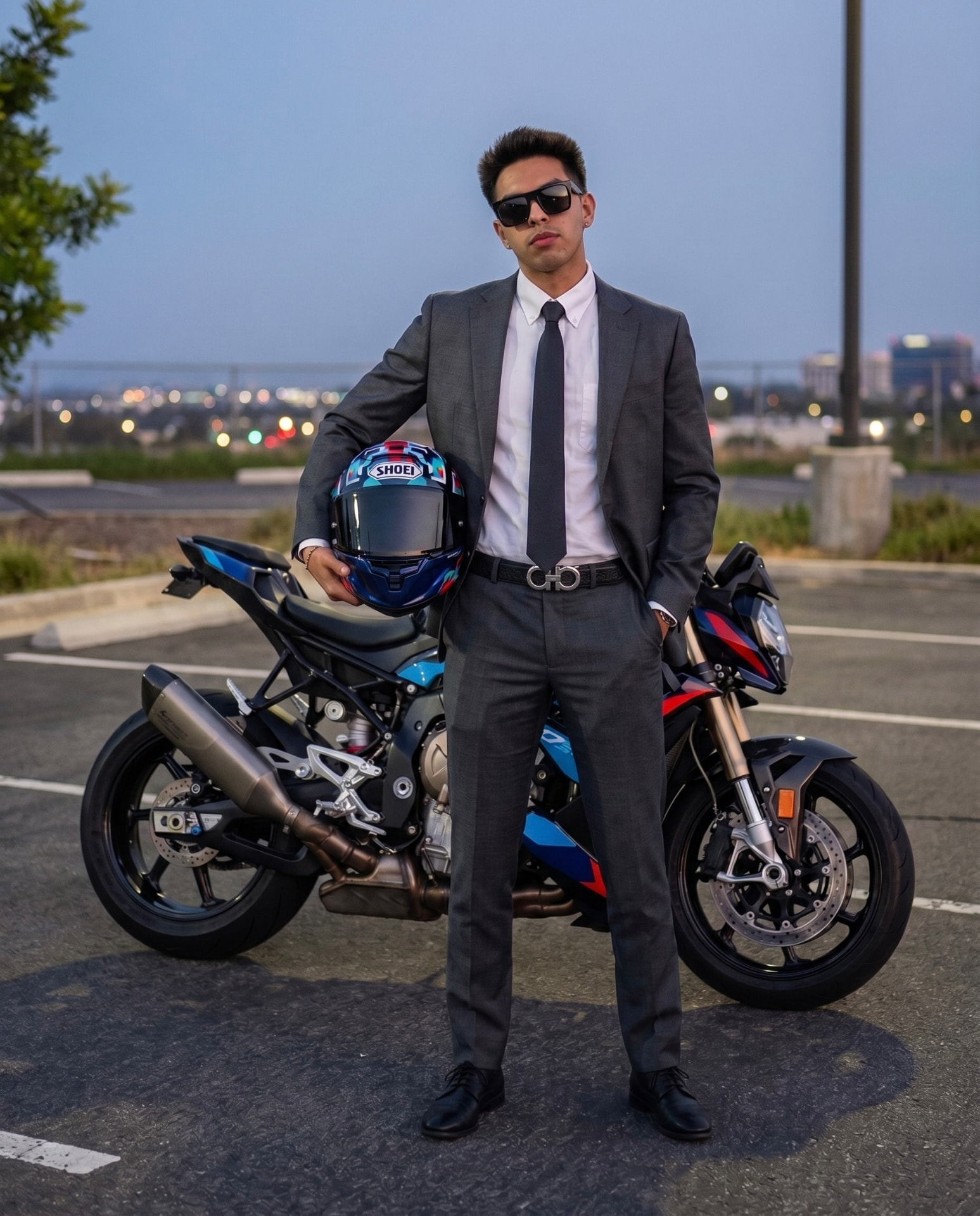 Rider in a charcoal suit holding a helmet beside a naked sport motorcycle at dusk